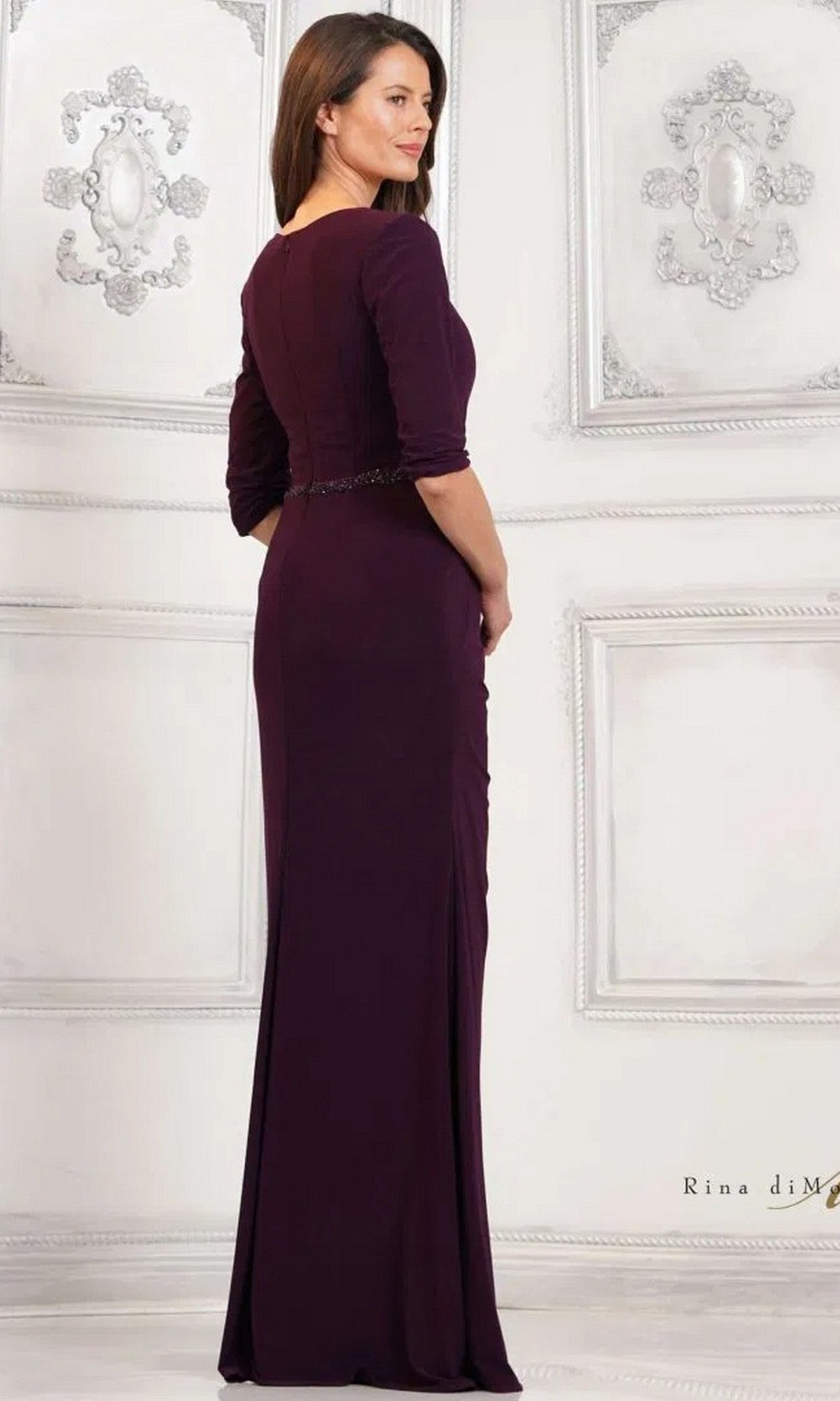 A woman stands in an ornate white room, wearing the Rina Di Montella RD3109 long formal dress in dark purple matte jersey with long sleeves and a beaded waistband, facing away and looking over her shoulder.
